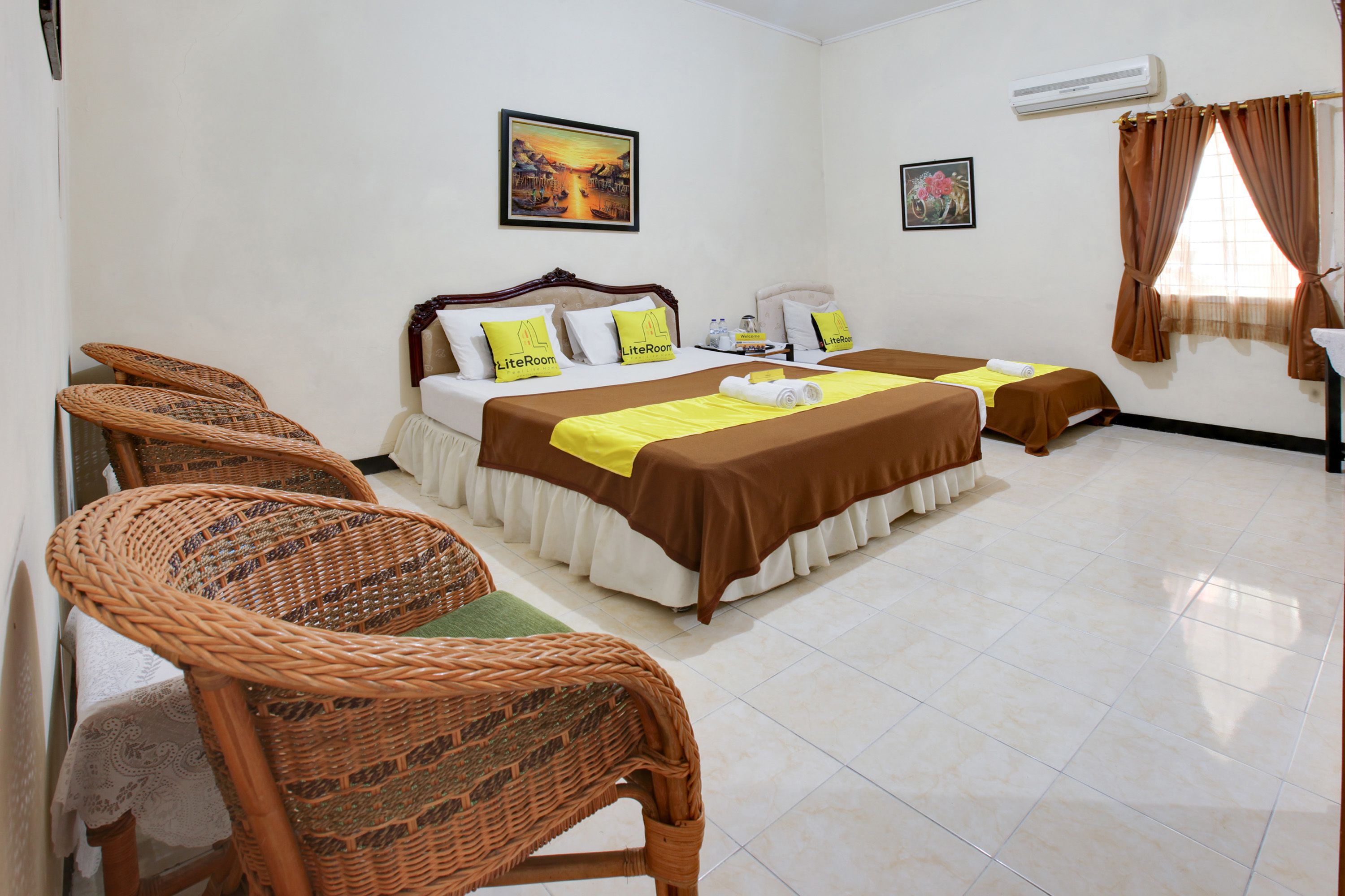 A hotel room with two beds, wicker chairs, a painting on the wall, and an air conditioner.