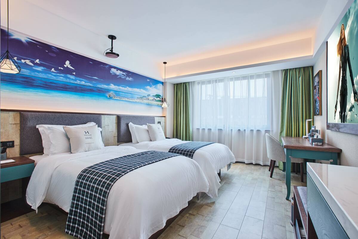Une chambre d’hôtel avec deux lits, un bureau, une chaise, une grande fresque murale et une fenêtre avec des rideaux.