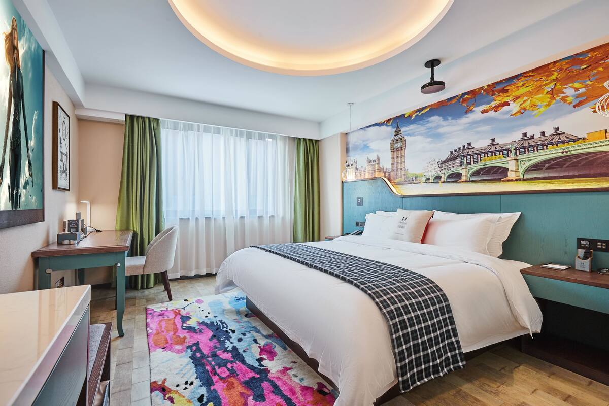Une chambre d’hôtel avec un grand lit, un bureau, une chaise, un tapis coloré et une fresque représentant un pont et une tour sur le mur.