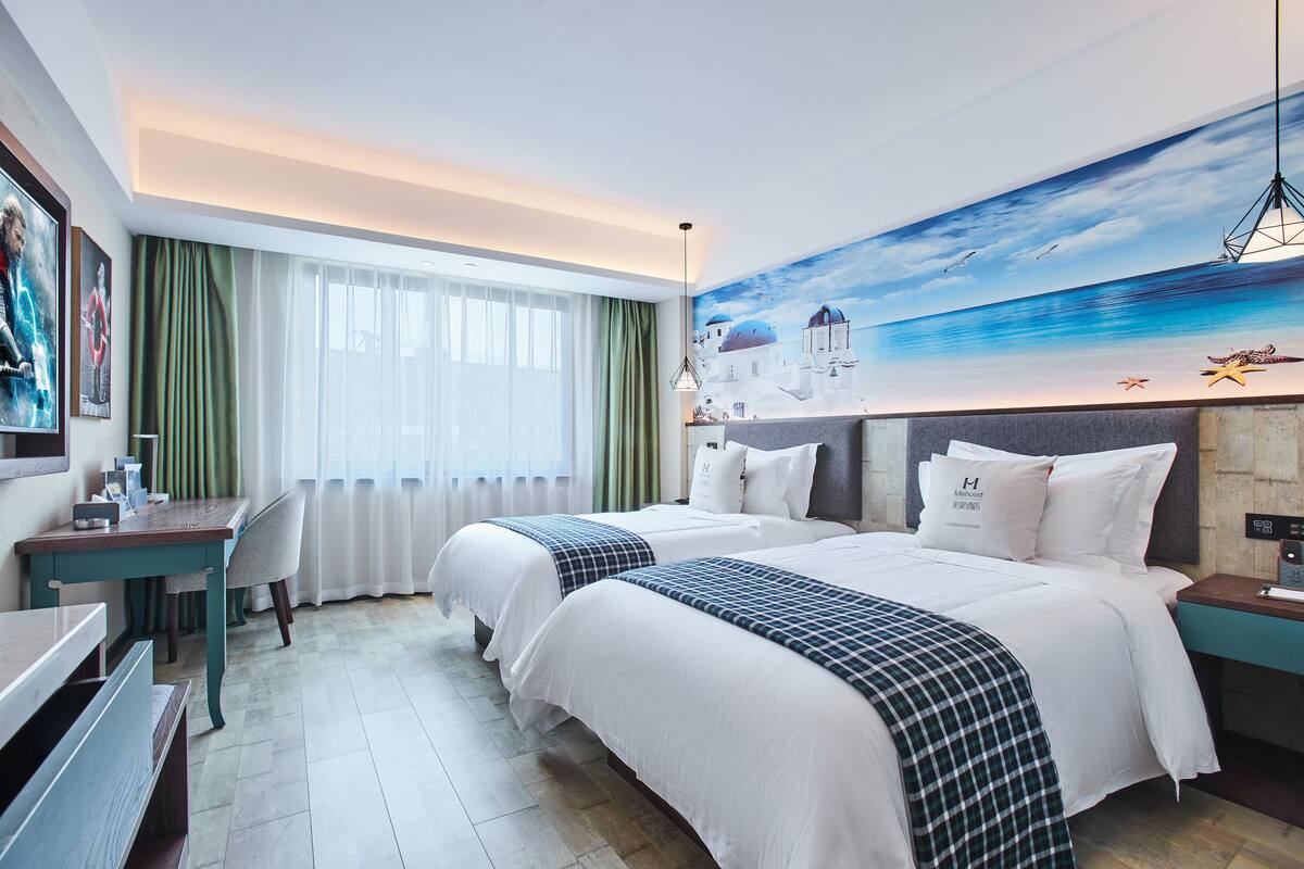 Une chambre d’hôtel avec deux lits, un bureau, une chaise et une grande fresque murale représentant un paysage côtier.