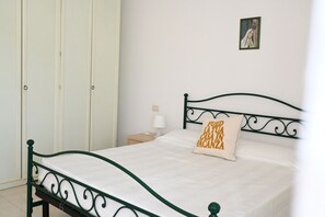 A single bed with a white mattress and a decorative pillow, a nightstand with a lamp, and a wardrobe with a door.