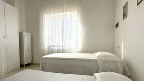 Ein Zimmer mit zwei Betten, einem Kleiderschrank, einer Kommode und einem Fenster mit durchsichtigen Vorhängen.