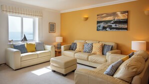 A living room with two beige sofas, a coffee table, and a window with a view of the sea. - Marine Point, Mevagissey - sensational cliff top views of harbour and bay (Mevagissey)