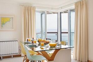 A modern dining area with a glass table, yellow chairs, breakfast items, and a view of the sea through large windows.