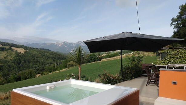 A hot tub with a wooden exterior, surrounded by a patio area with a table and chairs, and a view of a green landscape with mountains in the background. - A modern independent apartment with a large private terrace. (Macerata)