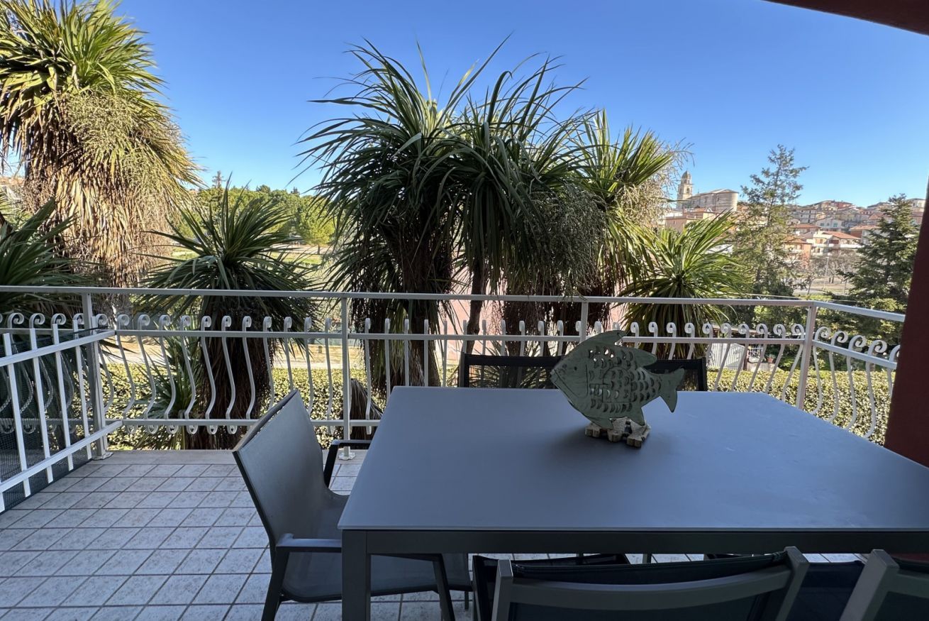 Un balcón con mesa y sillas, rodeado por una valla de metal blanco, con vistas a un paisaje urbano con edificios y palmeras.