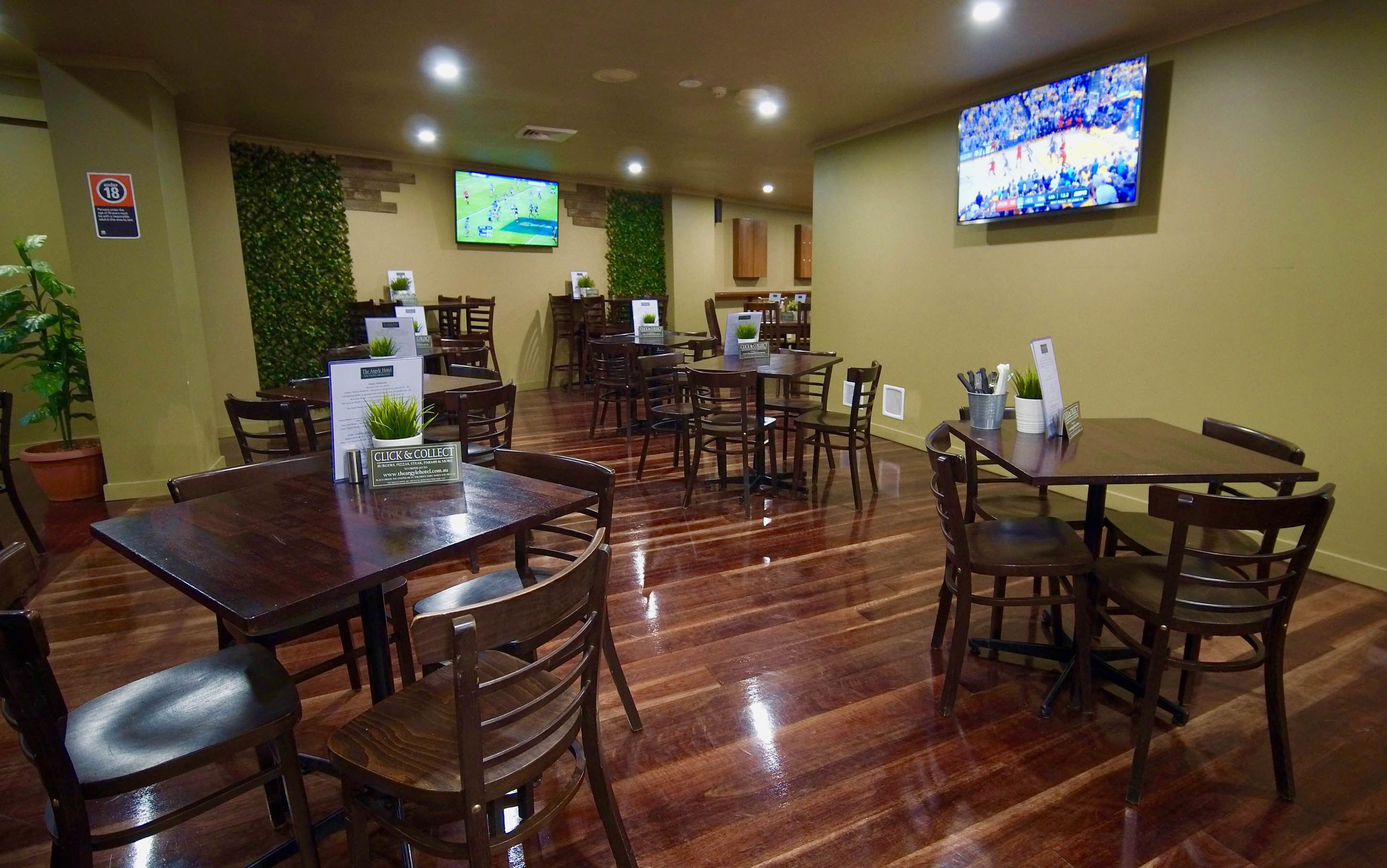A restaurant with wooden tables and chairs, a TV mounted on the wall, and a sign indicating a minimum age requirement.