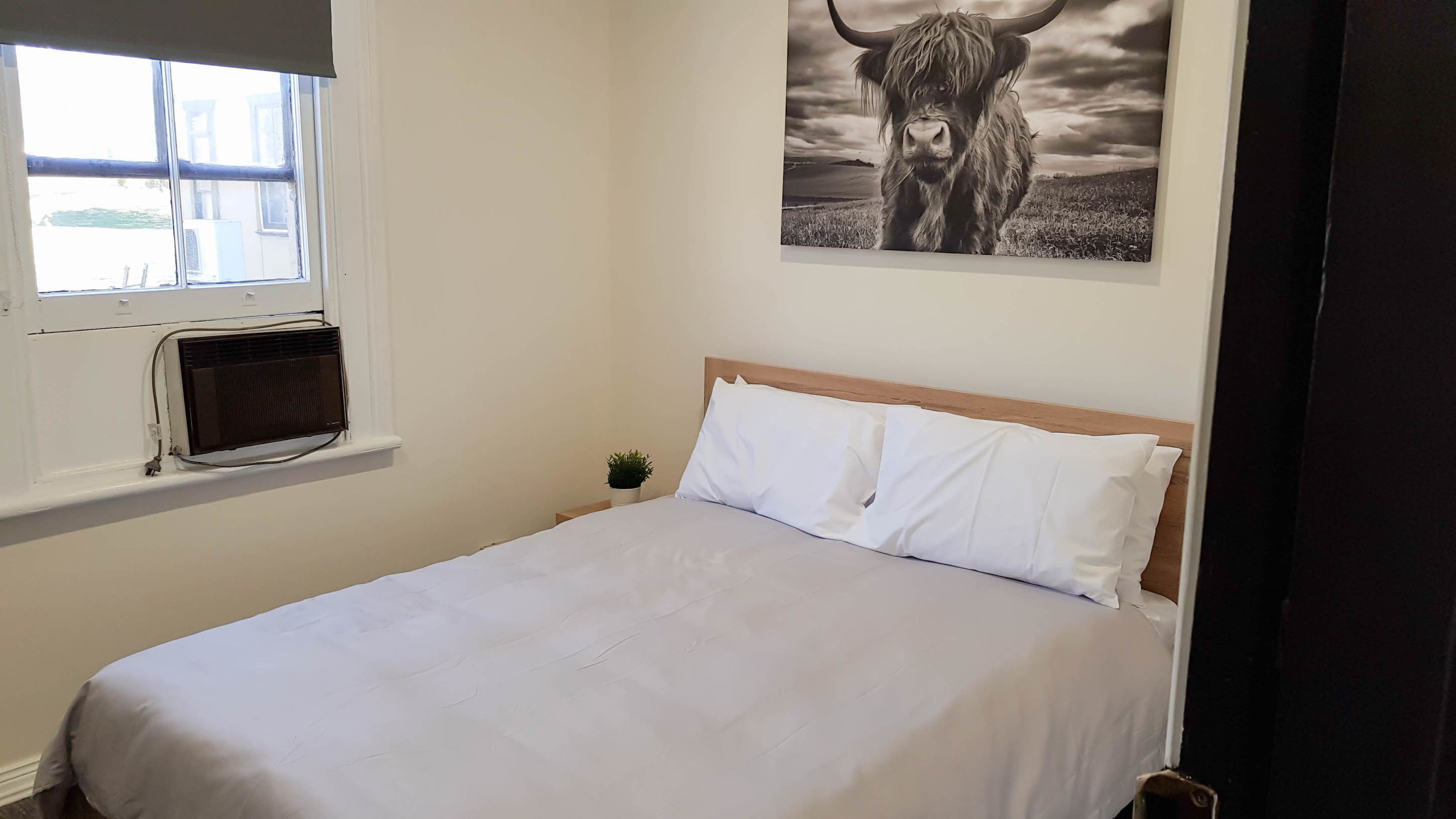 A bedroom with a bed, a window, an air conditioning unit, a small plant, and a painting of a bull.