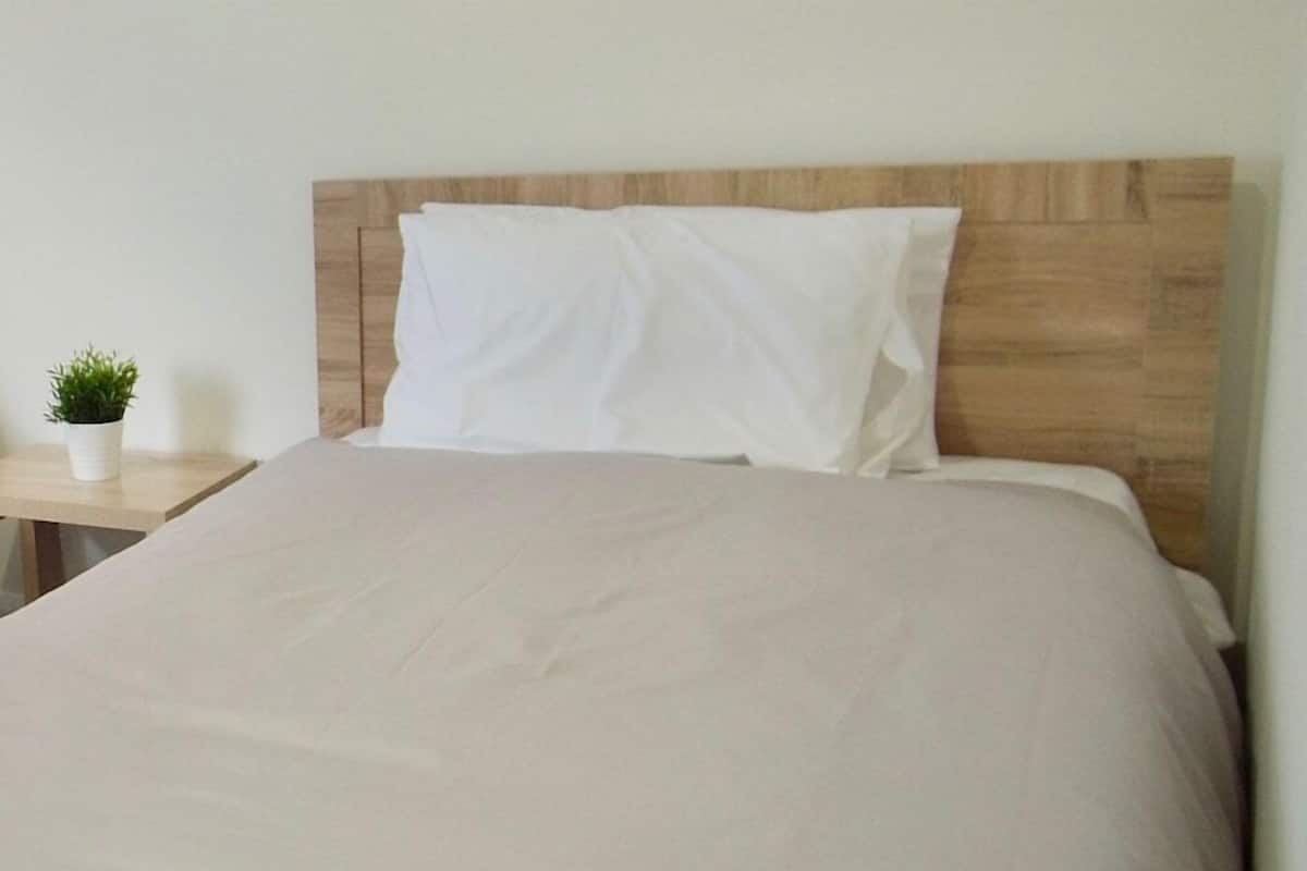 Una cama bien hecha con cabecera de madera, una mesita de noche con una planta en maceta y una pared lisa.