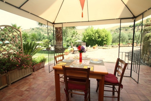 Une terrasse couverte avec une table dressée pour deux personnes, entourée de plantes en pot et offrant une vue sur la campagne.