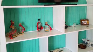 Una habitación con una pared color turquesa, un televisor de pantalla plana montado sobre una repisa blanca, y varios objetos en las repisas, incluyendo botellas, un teléfono y revistas.