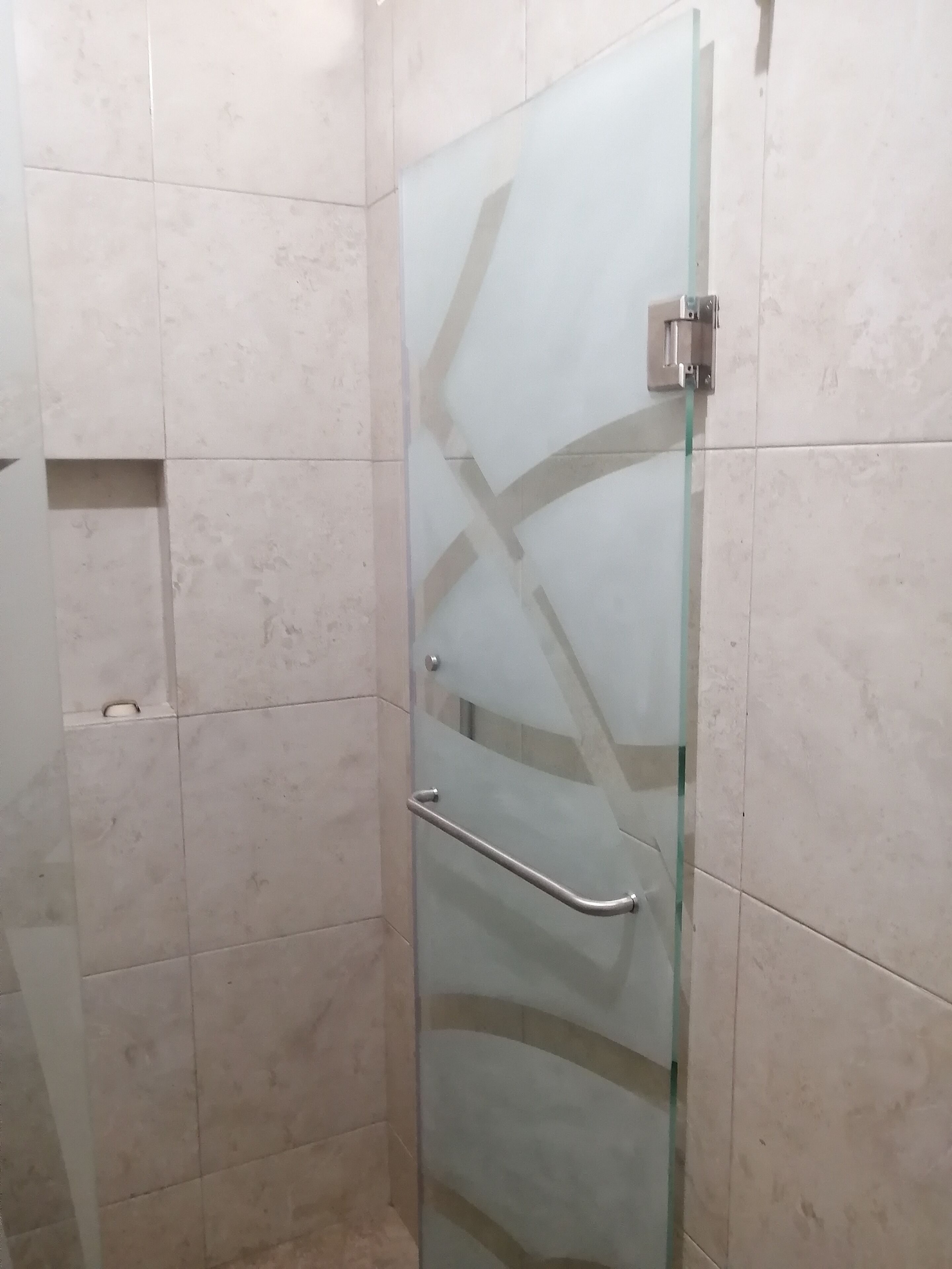 Cabina de ducha con paredes de azulejos, una barra de apoyo metálica y desagüe visible, todo ello envuelto en cristal.