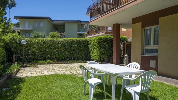 A grassy outdoor area with a white table and chairs, a hedge, and a building with balconies. - M152 - Marcelli, new three-room apartment with garden (Marcelli)