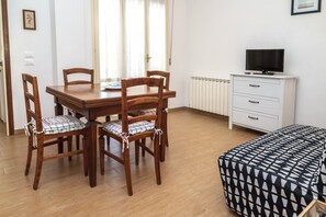 A room with a wooden dining table, four chairs, a white dresser with a TV, and a patterned bed.