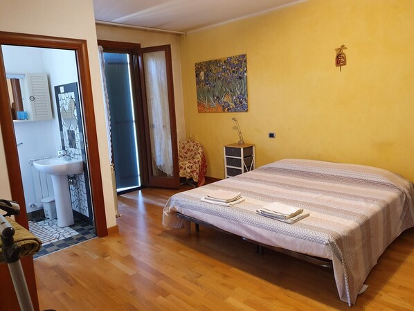 A bedroom with a bed, a bedside table, a chair, a painting, and a bathroom visible through an open door.