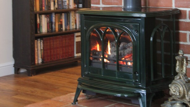 A room with a brick wall, a wooden bookshelf filled with books, and a green wood-burning stove. - 3 Bedroom historic house in Norfolk (Thetford)