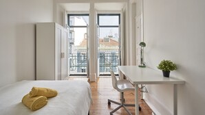Una habitación pequeña y luminosa con una cama, un escritorio y vistas a edificios a través de un ventanal grande.