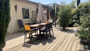 A wooden deck with a table and chairs, surrounded by greenery and a building with a tiled roof.