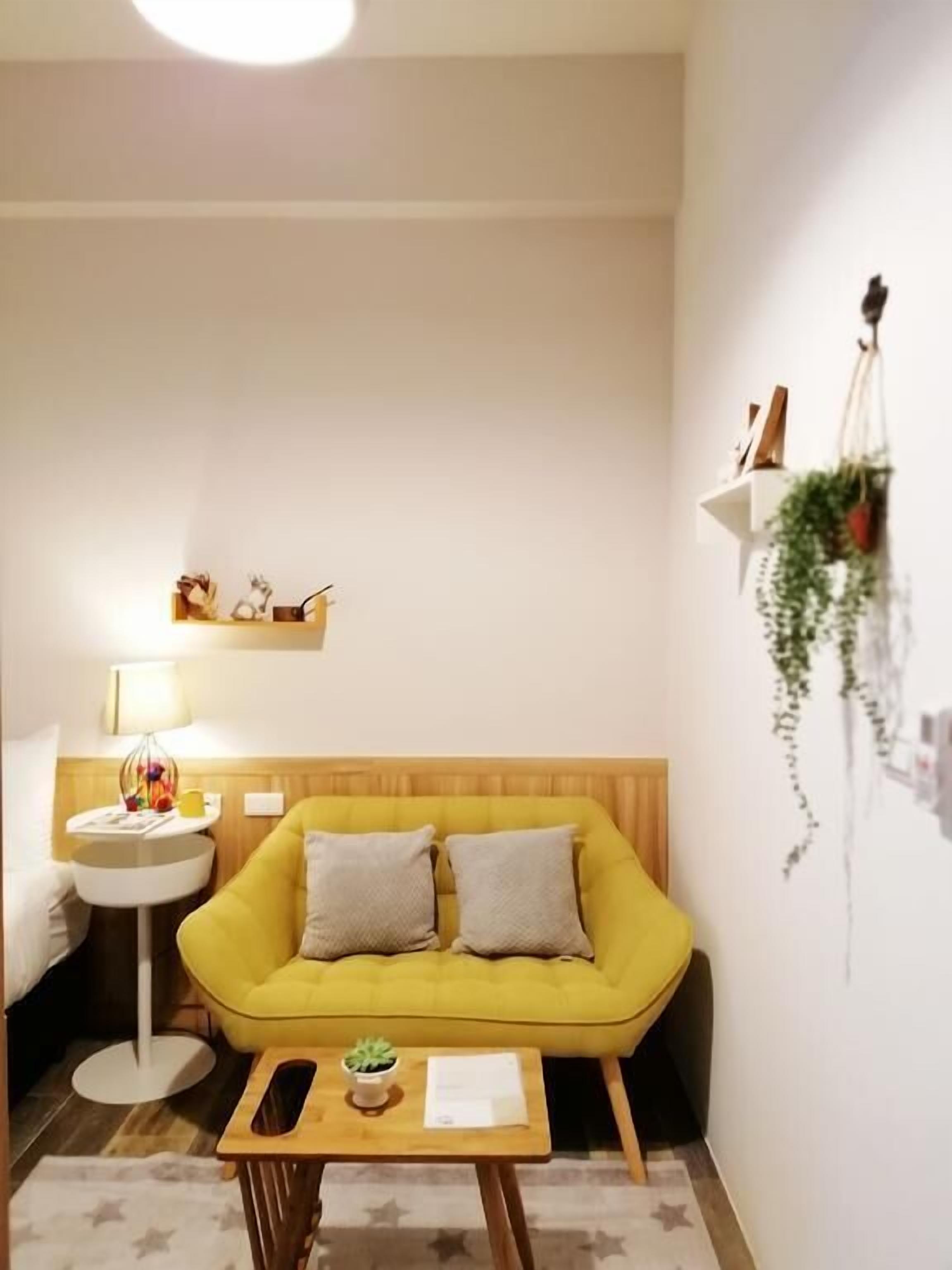 A yellow sofa with two pillows, a small wooden table with a plant and a book, a white side table with a lamp, and a wall-mounted shelf with decorative items.