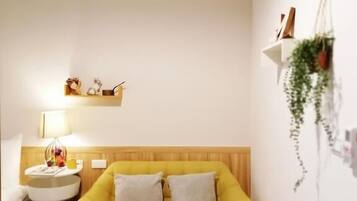 A yellow sofa with two pillows, a small wooden table with a plant and a book, a white side table with a lamp, and a wall-mounted shelf with decorative items.
