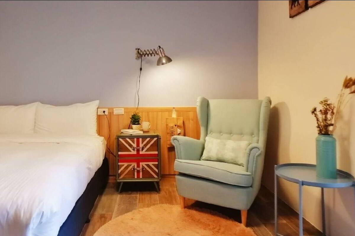 A bedroom with a bed, a chair, a nightstand with a Union Jack flag, a lamp, and a small table with a vase.