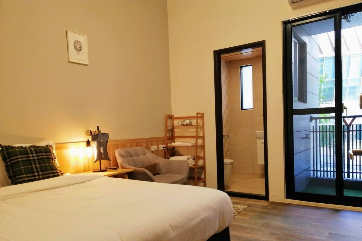 A bedroom with a bed, a chair, a small table, a shelf with towels, and a sliding glass door leading to a balcony.