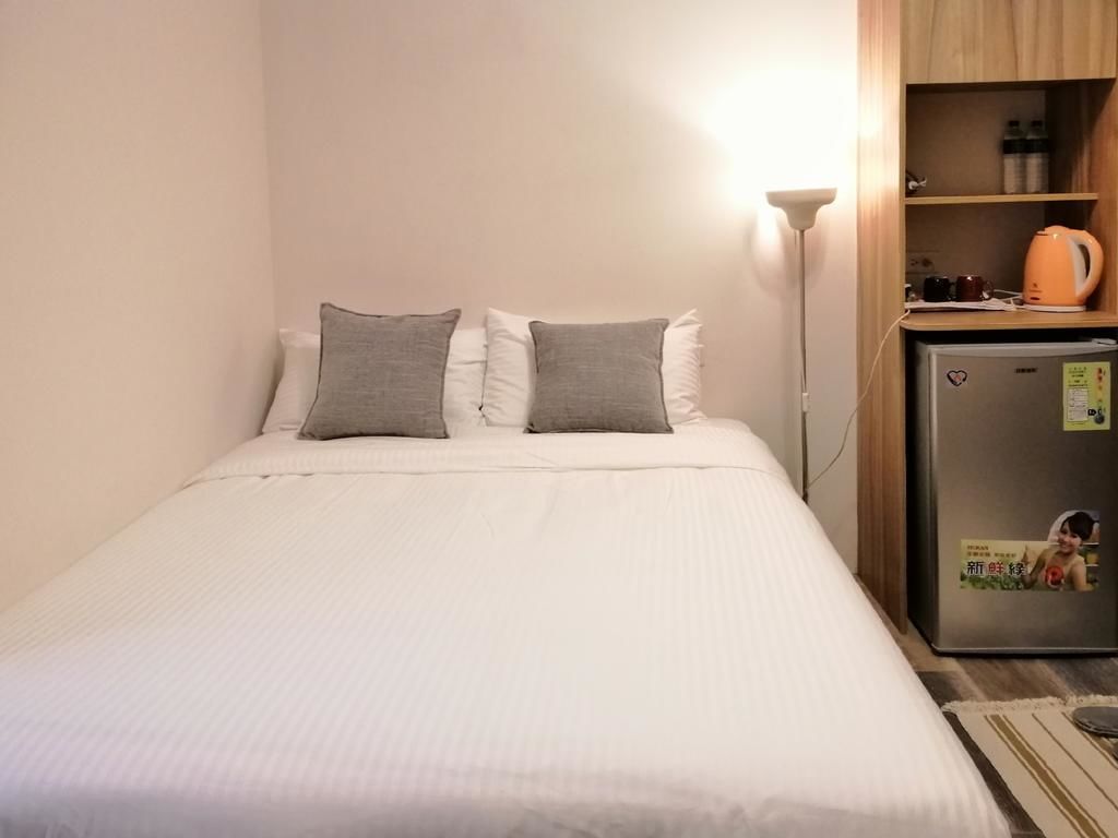 A neatly made bed with white bedding and two gray pillows, a small refrigerator with a sticker, and a lamp on a wooden shelf.