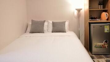 A neatly made bed with white bedding and two gray pillows, a small refrigerator with a sticker, and a lamp on a wooden shelf.
