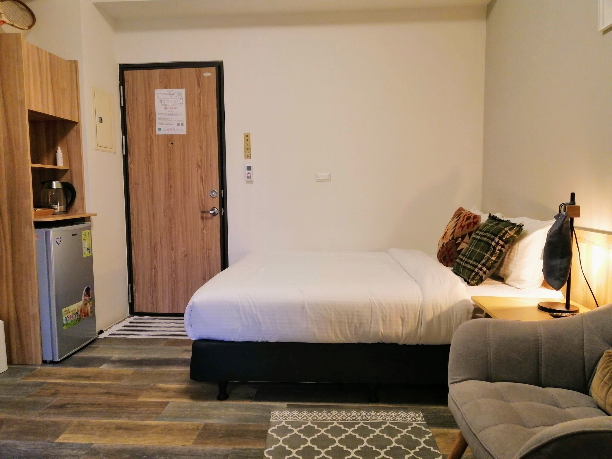 A compact hotel room with a bed, a sofa, a kitchenette, and a door with a notice.