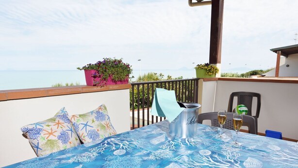 A balcony with a table set for two, featuring a tablecloth with a marine pattern, a metal bucket, two glasses, and a lemon. There are potted plants on the railing and a chair with a green bag.