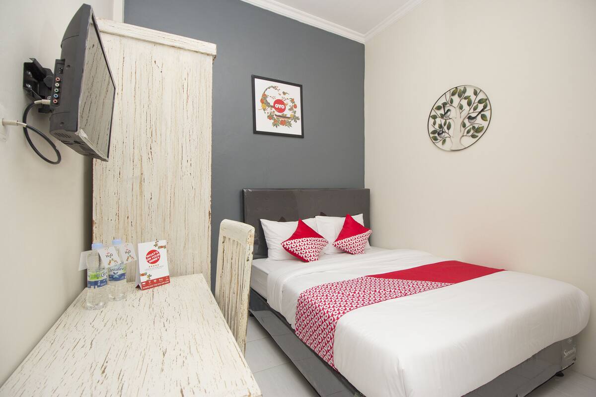 Habitación de hotel con cama, televisión montada en la pared y una mesita con botellas de agua y una tarjeta.
