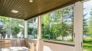 A covered patio with a table and chairs, large windows offering a view of trees and a road.