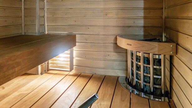 A sauna room with wooden walls and floor, a bench, and a steam room with a metal grate.