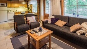 A cozy living room with a sofa, armchairs, and a coffee table. A kitchen is visible in the background with wooden cabinets and a microwave.