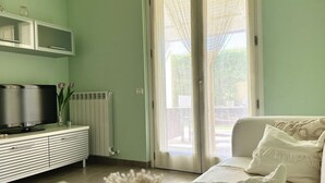 Ein Zimmer mit Fernseher auf einem weißen Schrank, einer weißen Couch und einer Schiebetür zum Außenbereich.