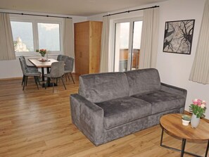 Una sala de estar moderna con un sofá gris, piso de madera, una mesa de comedor con sillas y vistas amplias a través de grandes ventanales.