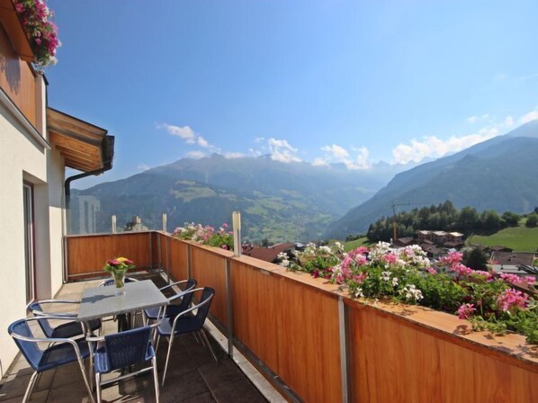 Un balcón con mesa y sillas, con vistas a un paisaje montañoso con casas y vegetación.