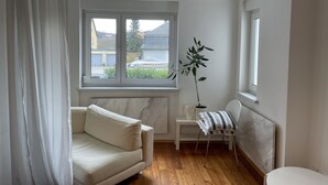 Une pièce avec un canapé, une petite table, une plante en pot et une fenêtre donnant sur les maisons à l’extérieur.