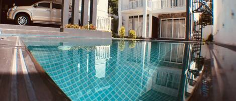 Une piscine à l’eau d’un bleu limpide, entourée de bâtiments modernes dotés de balcons, avec une voiture garée à proximité.