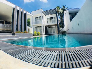 Un espace piscine moderne avec une piscine à l’eau d’un bleu limpide, un coin salon abrité et un escalier menant à un deuxième étage.