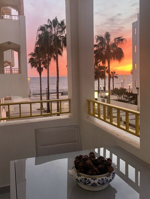 A balcony with a view of the sunset over the ocean, palm trees, and a table with a decorative bowl.