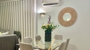 Un comedor moderno con una mesa de vidrio rodeada por cuatro sillas, un jarrón decorativo con flores, un aire acondicionado montado en la pared y un espejo redondo.