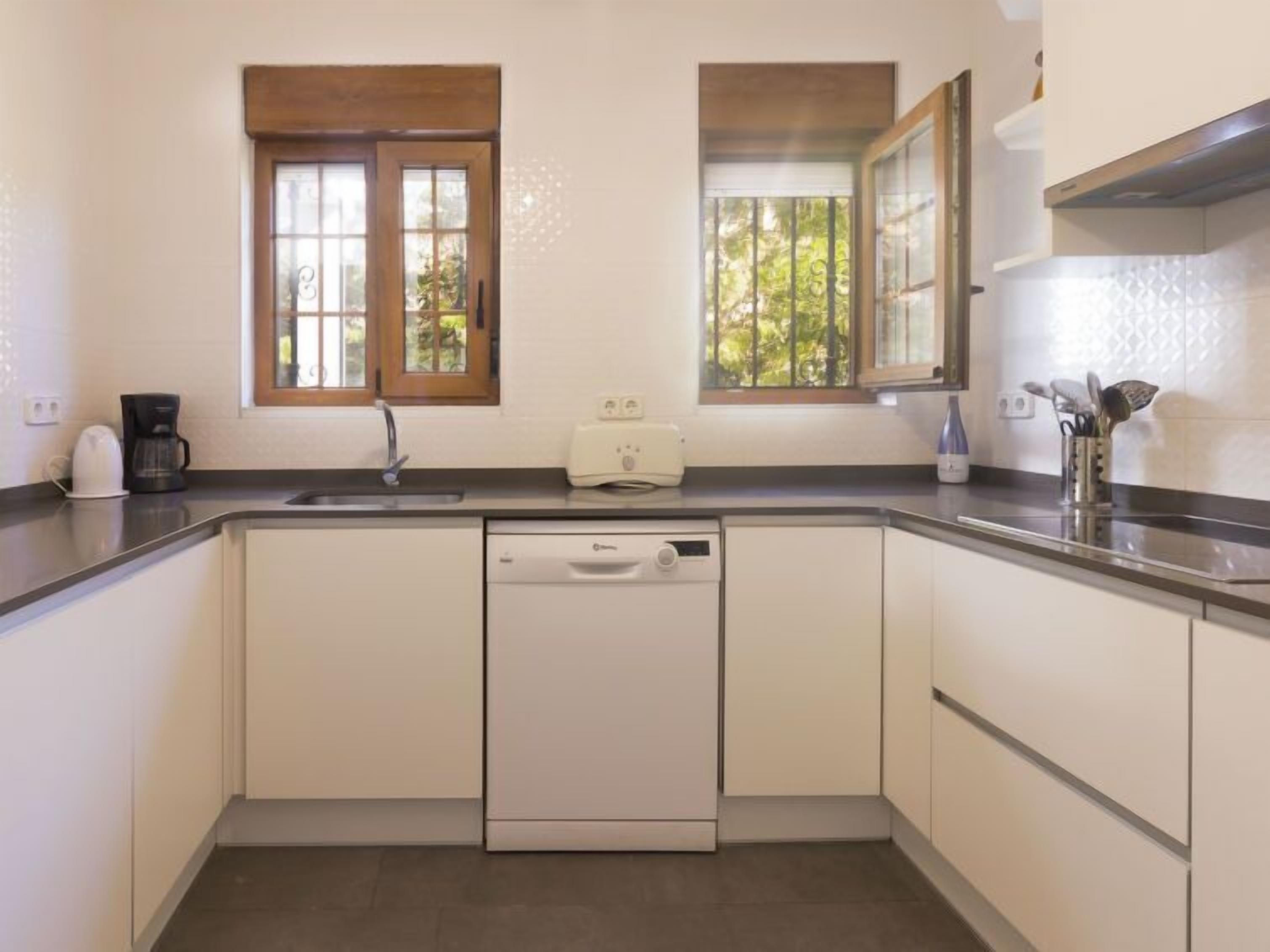 Cocina compacta equipada con lavavajillas, cafetera y tostadora. Dos ventanas ofrecen luz natural y vistas al exterior con vegetación.