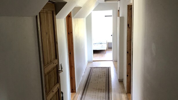 A narrow hallway with wooden flooring and a central rug, leading to a bedroom with a bed and a window. - In nature, close to the city (Aabenraa)