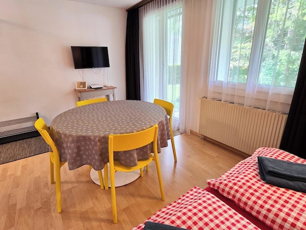 A room with a round table, yellow chairs, a flat-screen TV, a radiator, and a window with curtains.