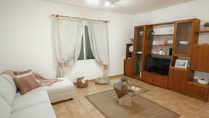 A living room with a white sofa, a glass coffee table, a wooden TV stand, and a window with white curtains. - Slowing Villas - Villa Oceano (Porto Santo)