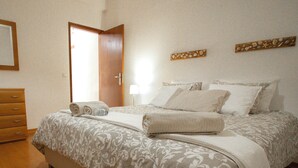 A neatly made bed with a patterned bedspread and pillows, a wooden dresser to the left, and a door leading to another room. - Slowing Villas - Villa Oceano (Porto Santo)