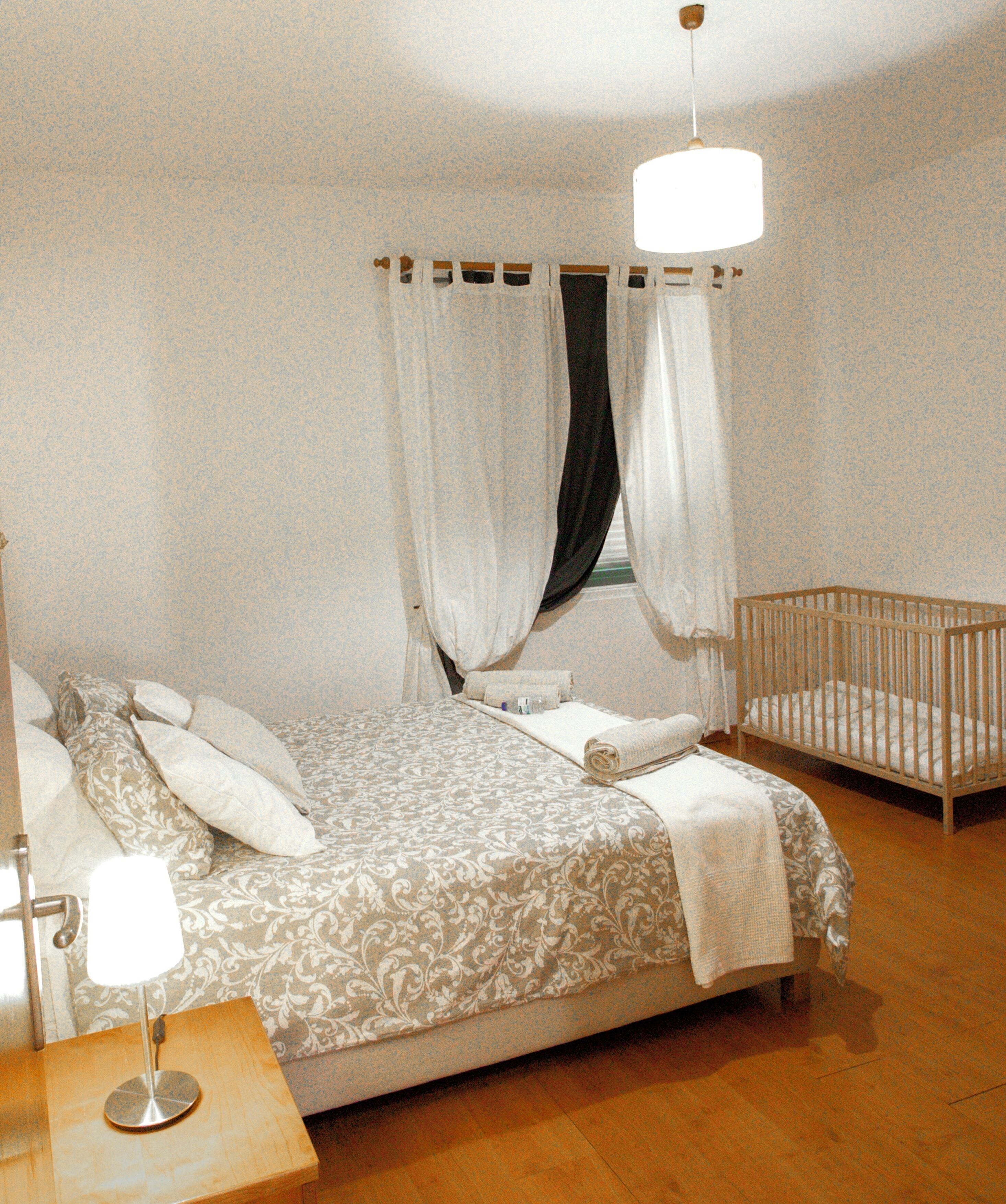 Un dormitorio con cama, cuna, mesita de noche y ventana con cortinas.