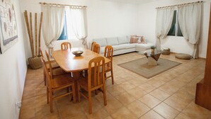 A dining area with a wooden table and chairs, a sofa, and a window with curtains. - Slowing Villas - Villa Oceano (Porto Santo)