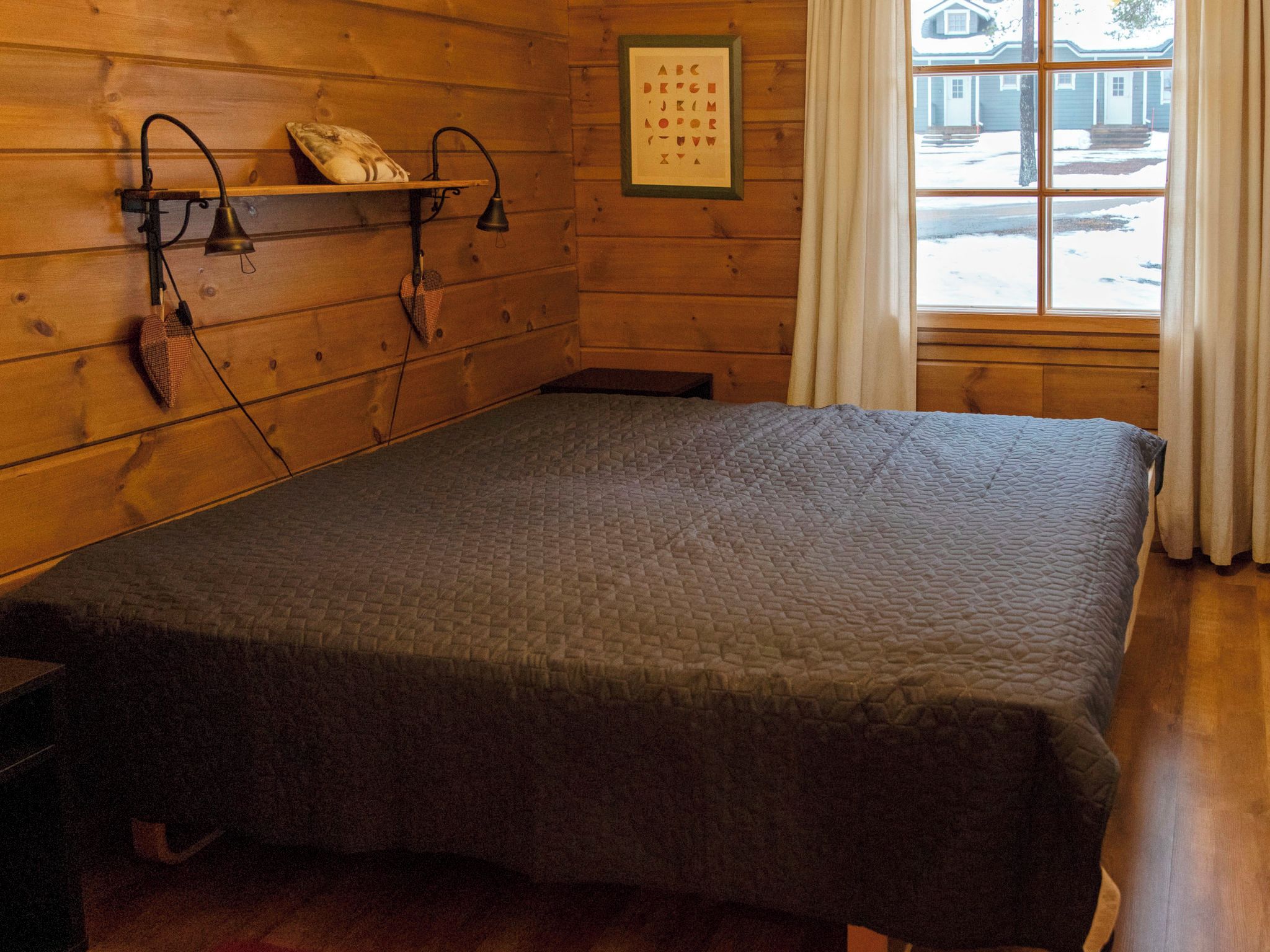 A wooden room with a bed, a wall-mounted shelf with a pillow, a framed alphabet chart, and a window with curtains.